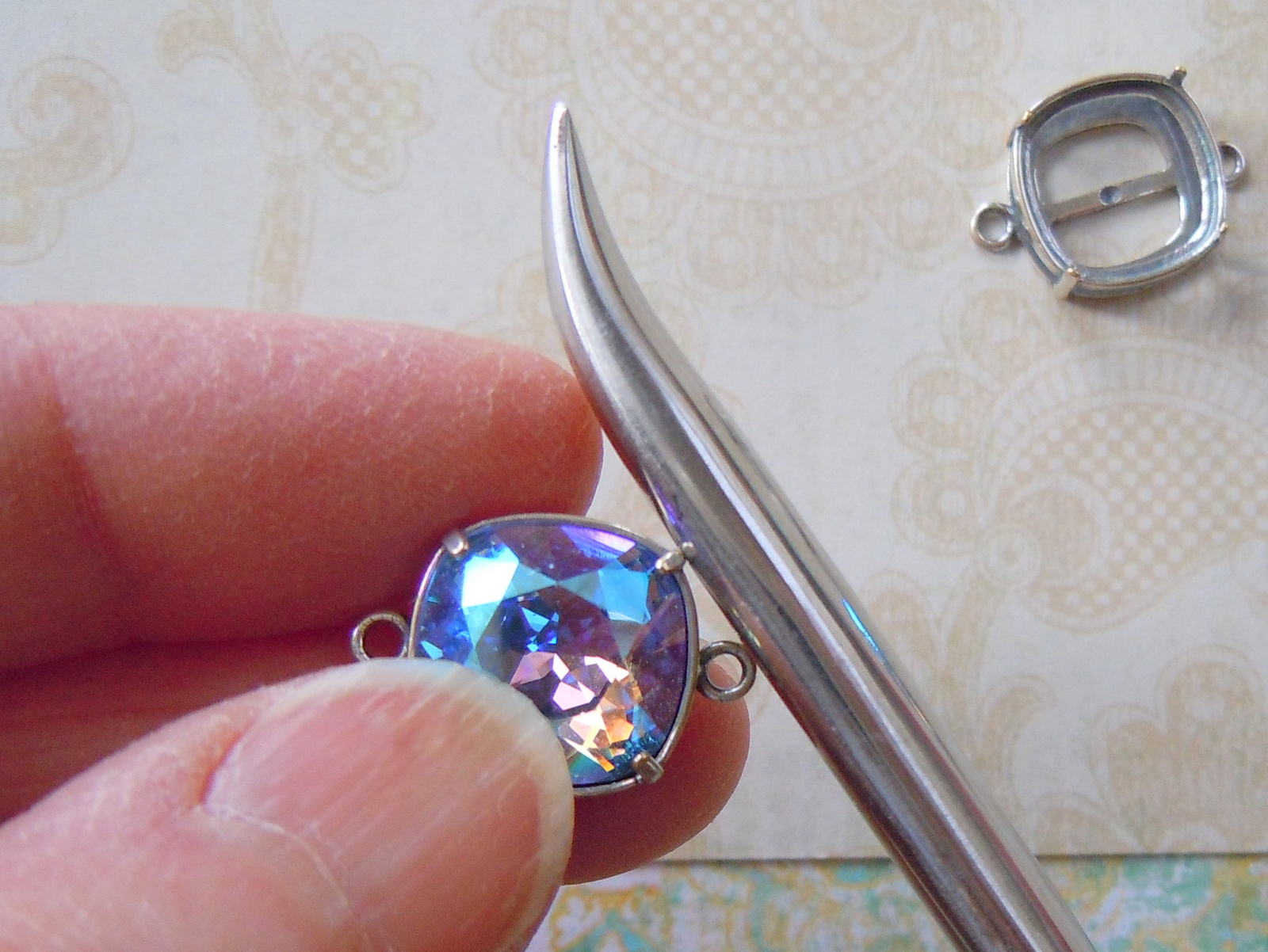 Place a Swarovski square cushion stone in the bezel setting. Firmly hold the bezel setting (or work on a rubber block). Use the burnisher to carefully push the first bezel prong until it is set against the stone. Rotate the bezel setting so the second prong you set is opposite the first one. Then set the final two prongs in the same manner. Repeat these steps with the second stone and setting. 